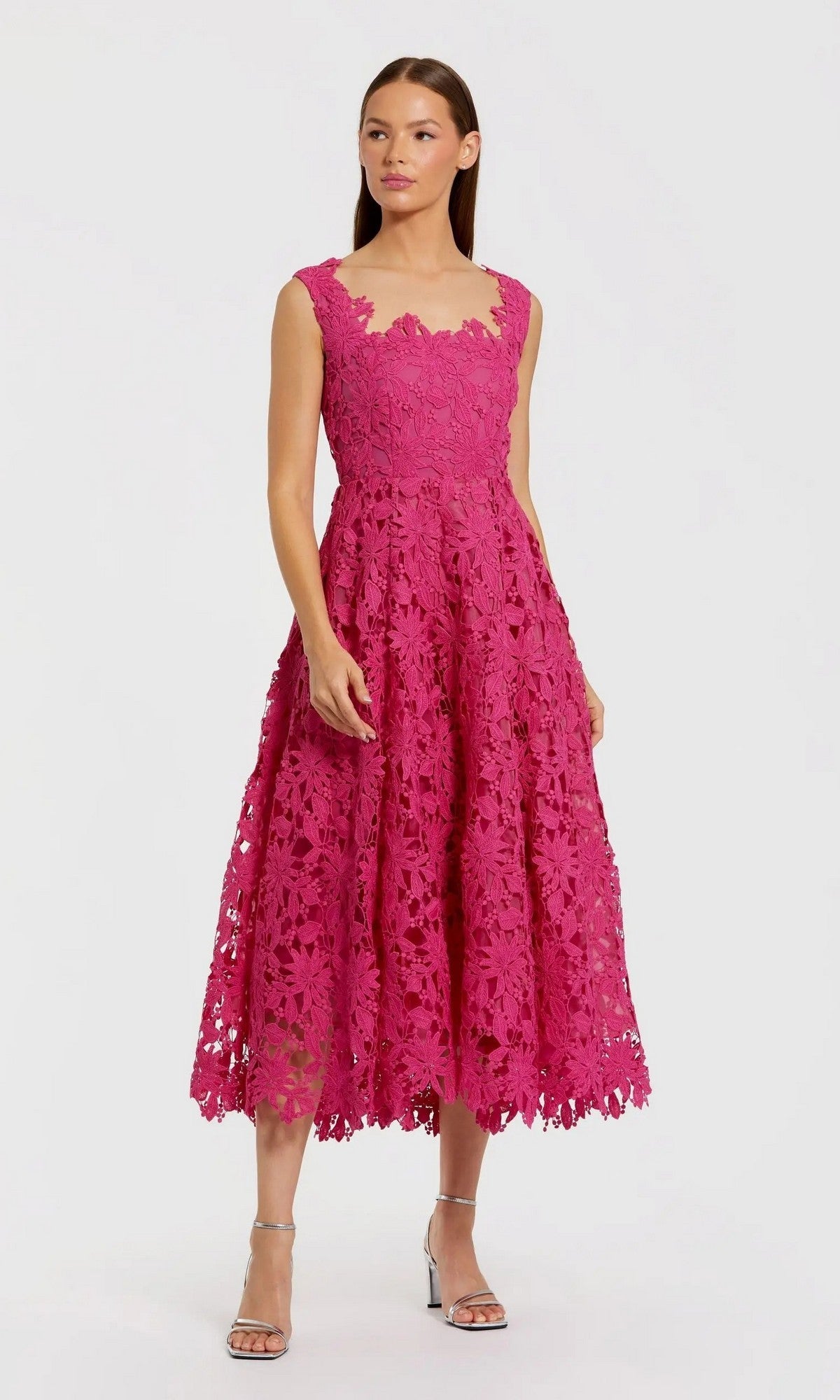 A woman wearing the Mac Duggal 69073 Midi Party Dress, featuring a sleeveless pink floral lace design with a fitted bodice and flared skirt, stands against a plain light background paired with silver high-heeled sandals.
